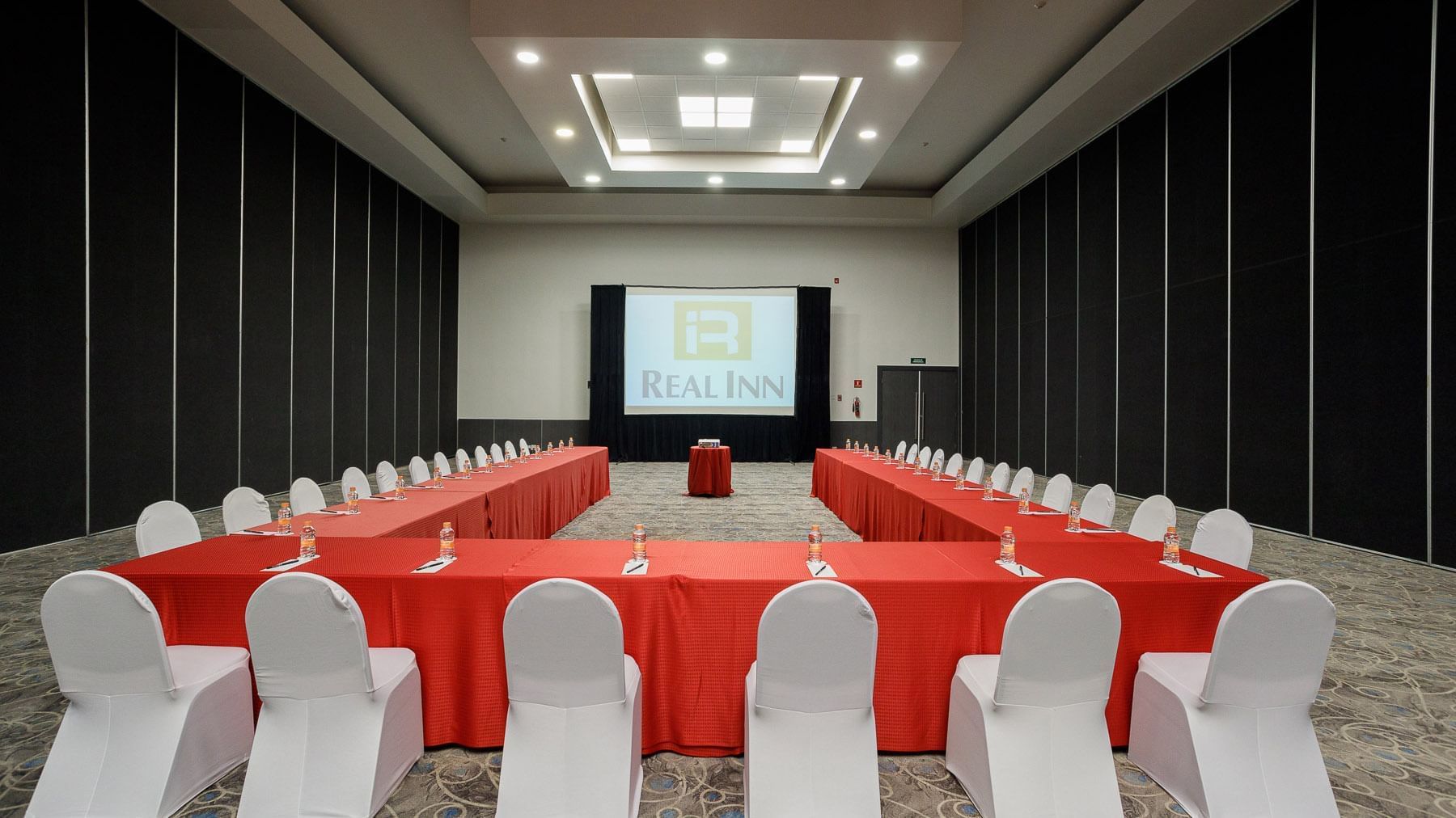 U-shaped meeting setup with red tablecloths and a large projector screen in Venecia Venue at Real Inn Tijuana