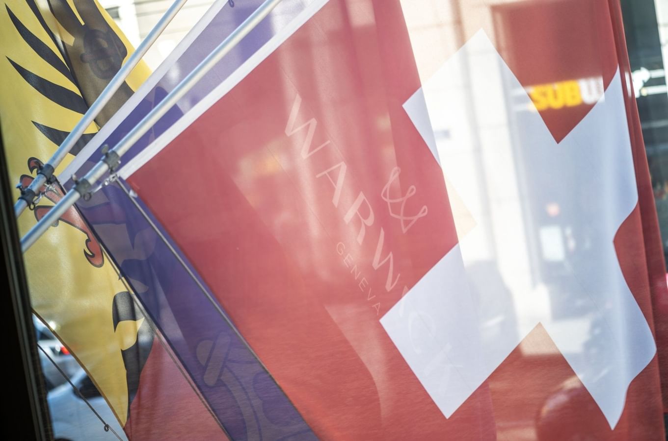 Drapeaux suisse et genevois avec le logo Warwick Geneva visible sur une façade vitrée au Warwick Geneva Genève