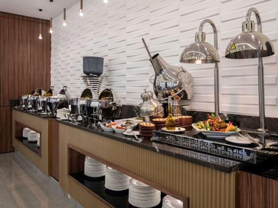 Buffet line with silver chafing dishes by a white textured wall in La Maison at Warwick Al Jubail