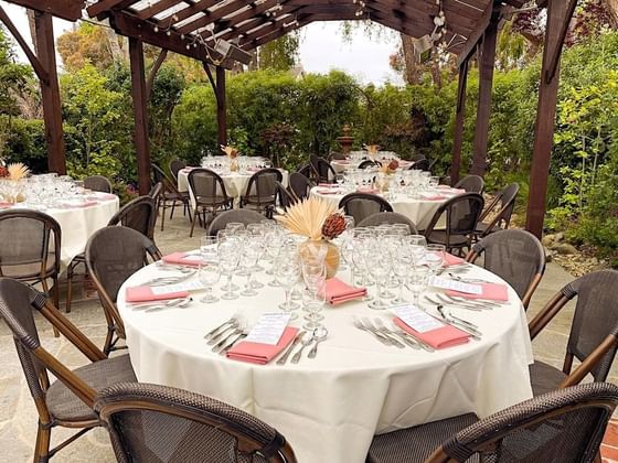 Outdoor dining area with round tables set with white linens and coral napkins under wooden pergola at Cambria Pines Lodge