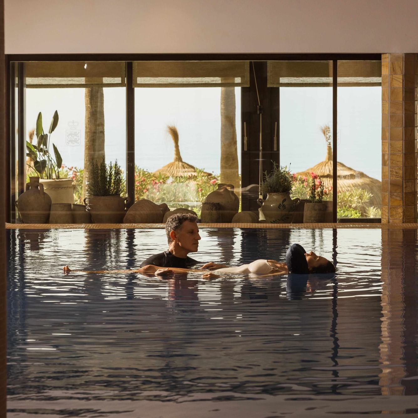 Pareja relajándose en piscina interior con vistas a sombrillas de paja y vegetación tropical al atardecer