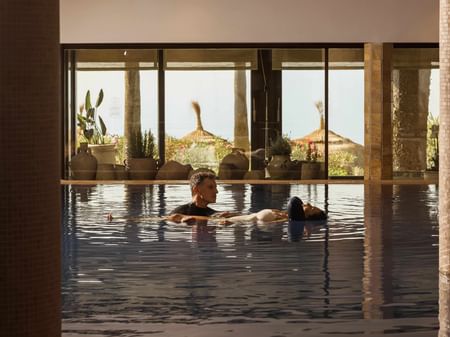 Pareja relajándose en piscina interior con vistas a sombrillas de paja y vegetación tropical al atardecer