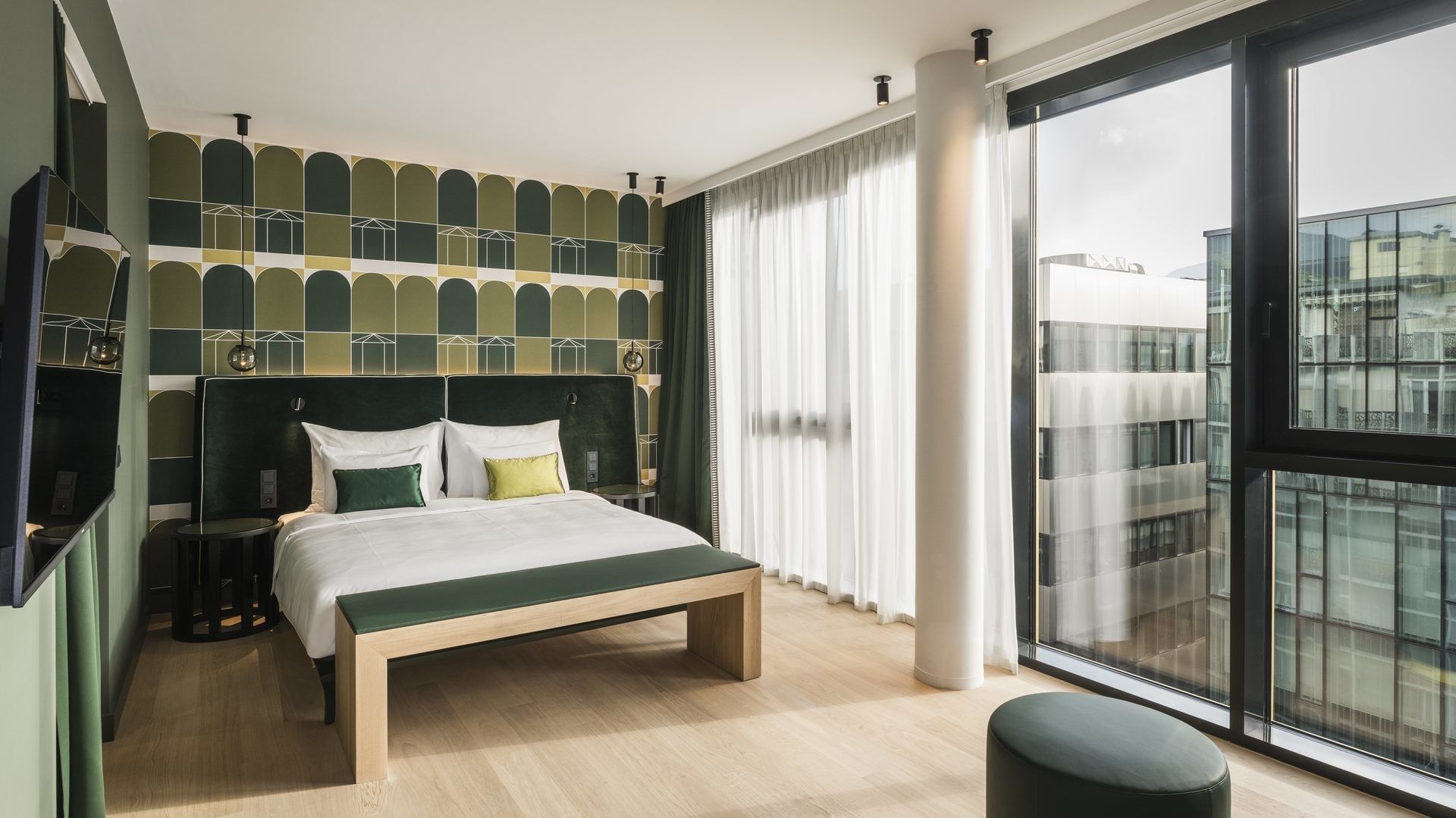 Modernes Hotelzimmer mit großem Bett, bunter Wand, großen Fenstern und Blick auf Gebäude.