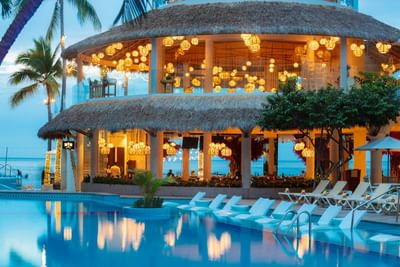Beachfront resort with thatched-roof building, illuminated lanterns, pool, and lounge chairs at Playa Los Arcos Puerto Vallarta