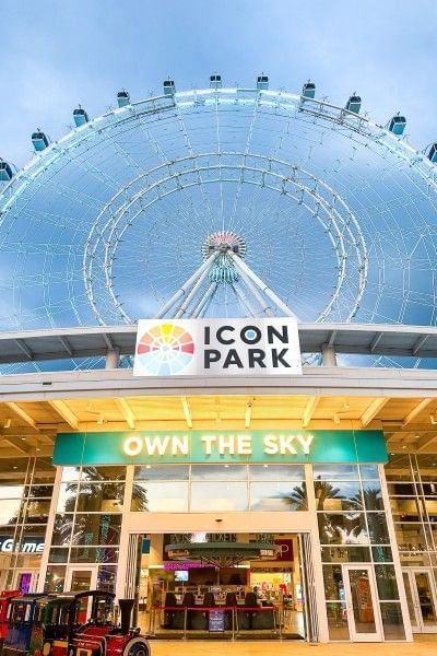 The entrance to a building with signs that read ICON Park and Own the Sky, with the Orlando Eye towering behind it against a darkening sky.