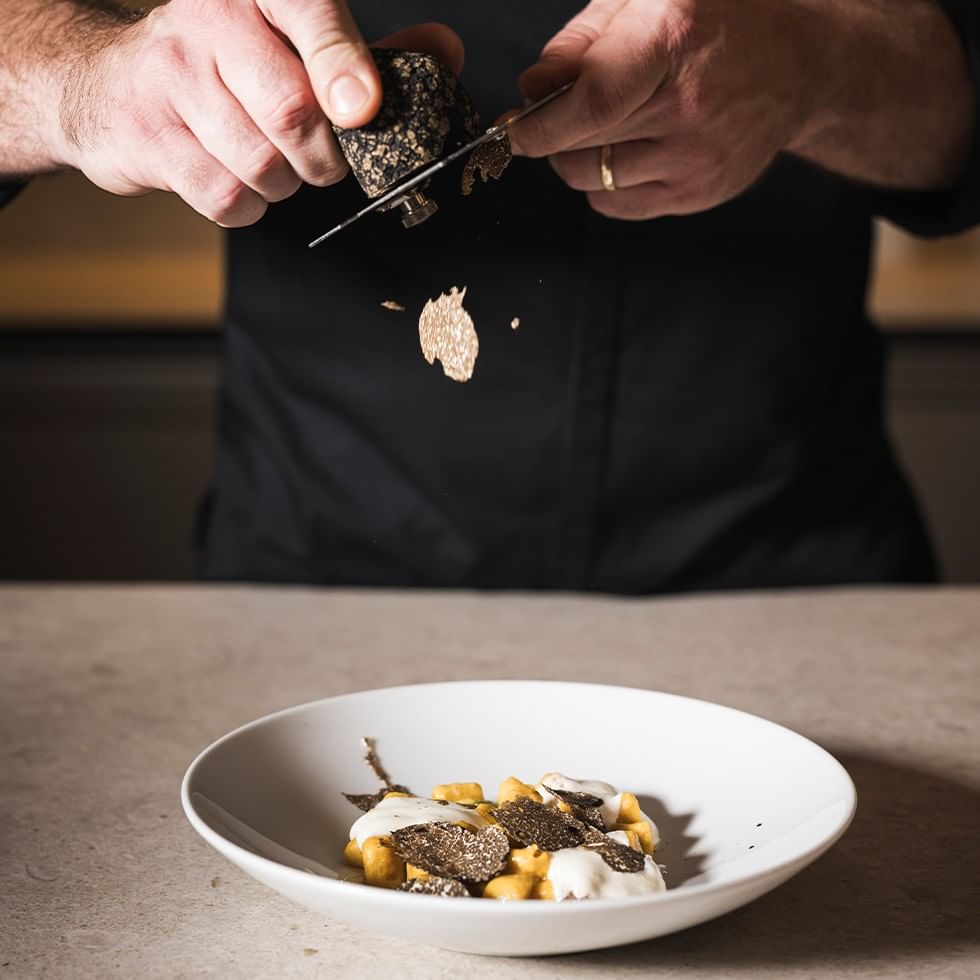 Chef in camicia nera che taglia tartufo nero su un piatto di gnocchi con tartufo.