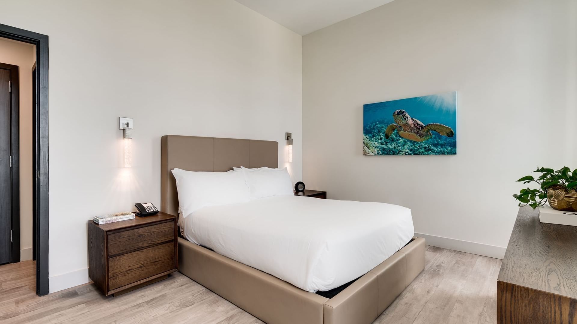 A neatly made bed with white linens and a brown leather headboard in a room with light wood floors and white walls.