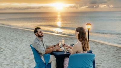 Couple in white outfits clinking champagne glasses on a beach near Amsterdam Manor Beach Resort