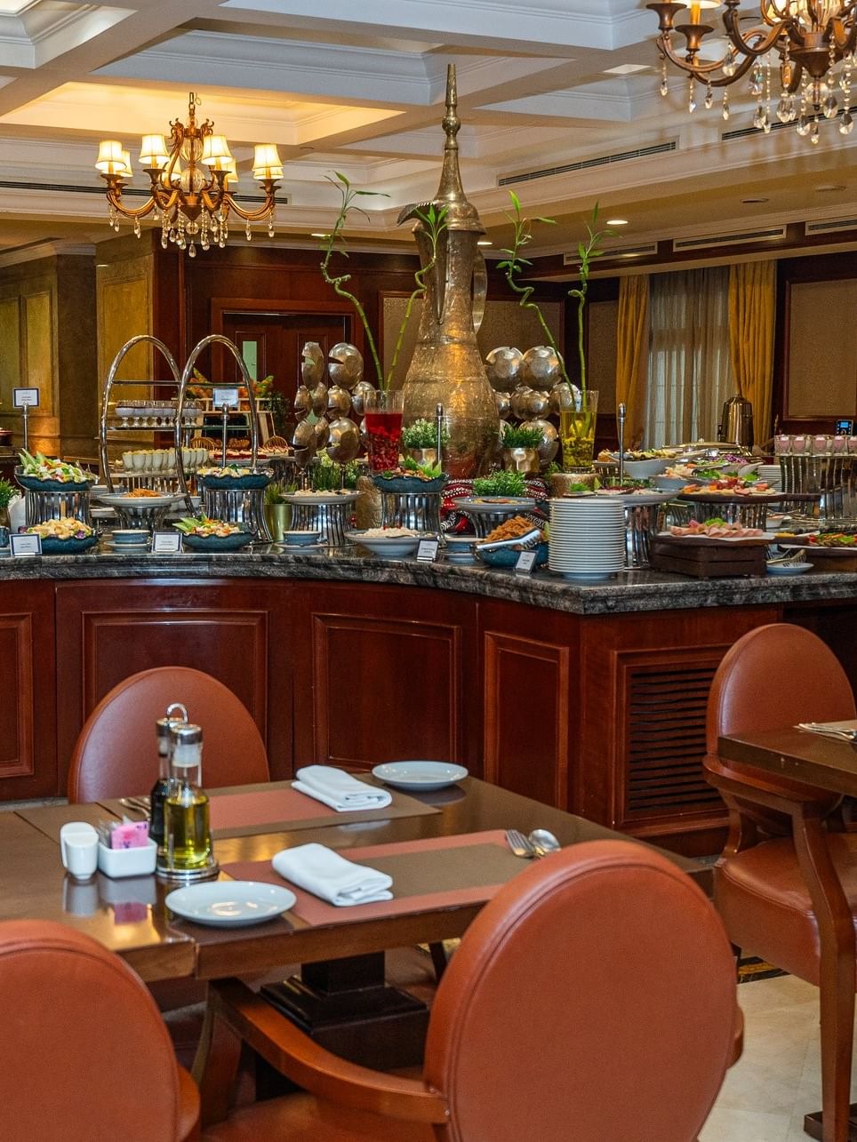 Dining tables by a curved buffet island under gold chandeliers in L’auberge at Warwick Doha Hotel