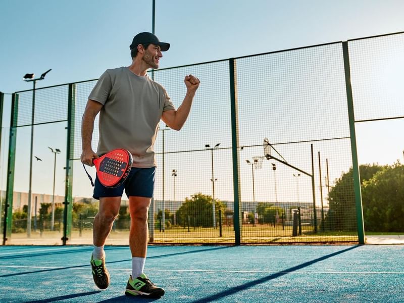 Uomo sorridente con racchetta da padel su campo sportivo, celebrando la vittoria con il pugno alzato.