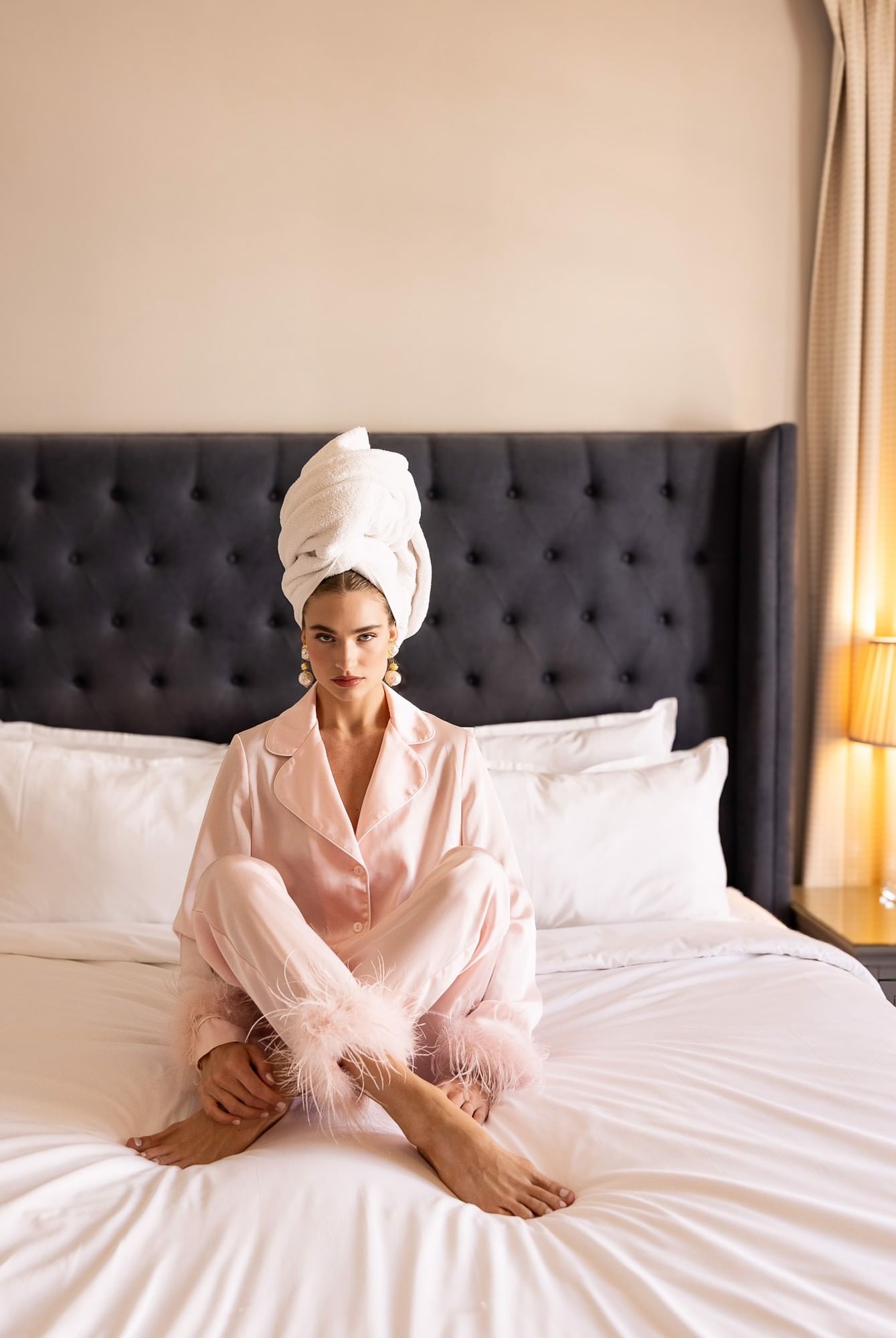 A woman in pink satin pajamas and a white towel on her head sits on a bed with a tufted headboard.