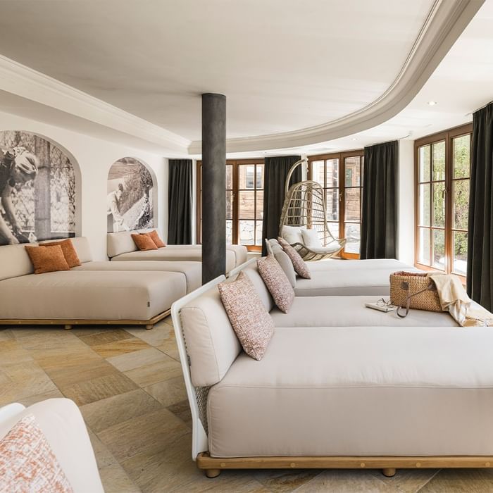 A room with three long couches, a hanging chair, large windows, and a wall with two arched images.