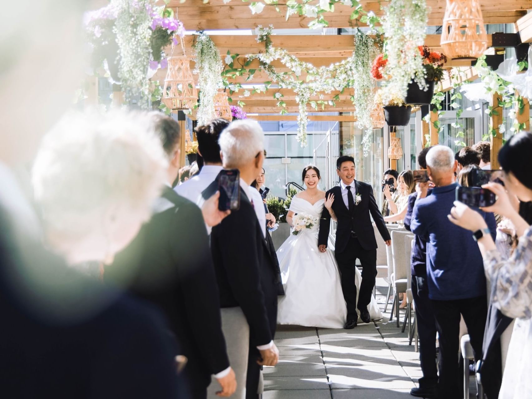 Bride walks with her father in a urban outdoor terrace with lots of guests