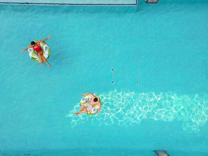Two guests on leaf-patterned floats by the edge of a bright blue swimming pool side at Warwick Melrose Dallas