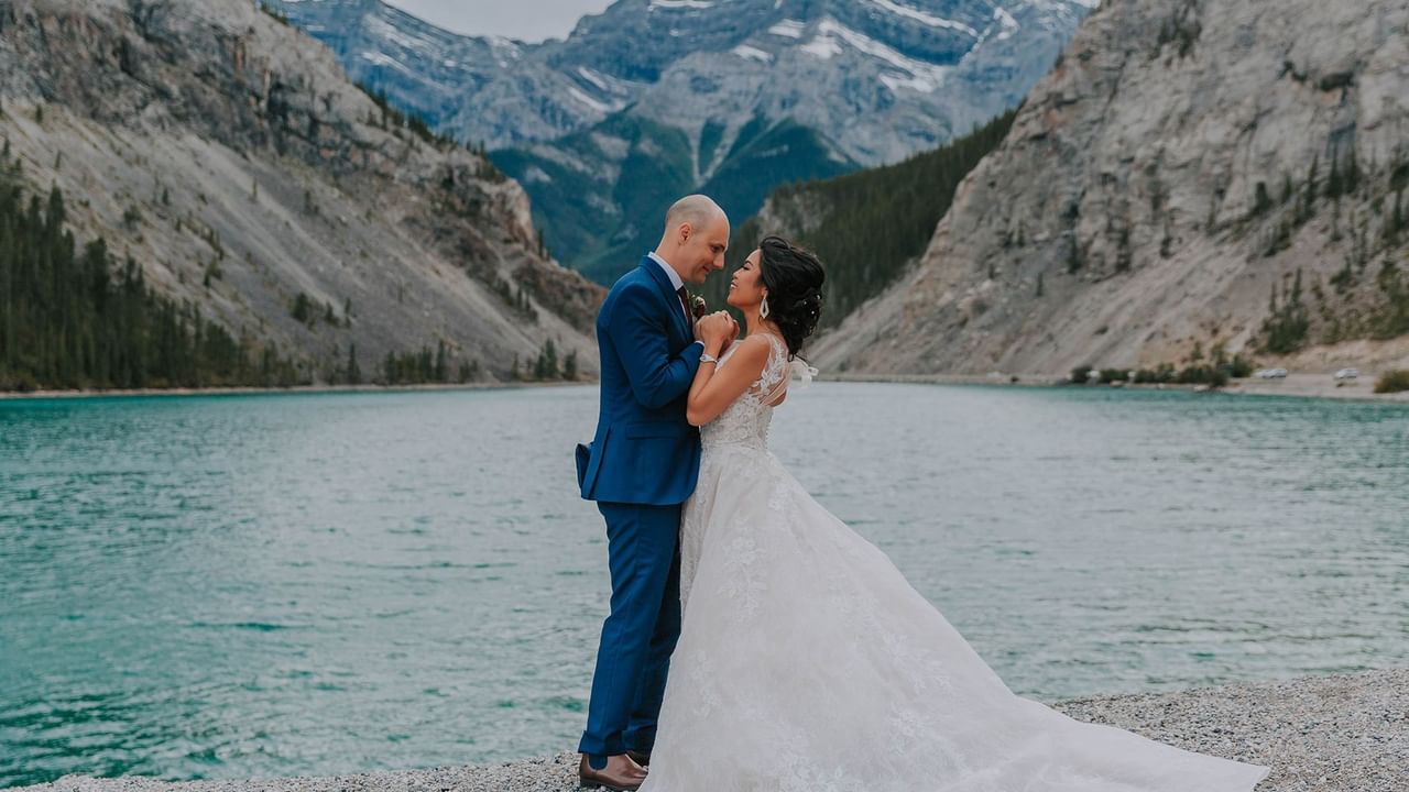 Bride and groom standing by a lake with mountains in the background.