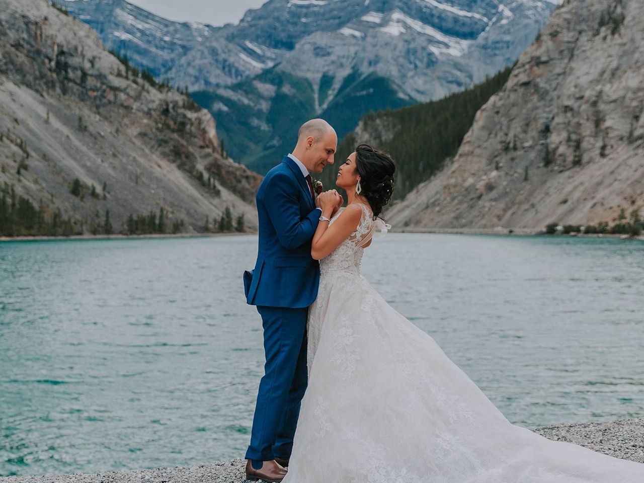 Bride and groom standing by a lake with mountains in the background.