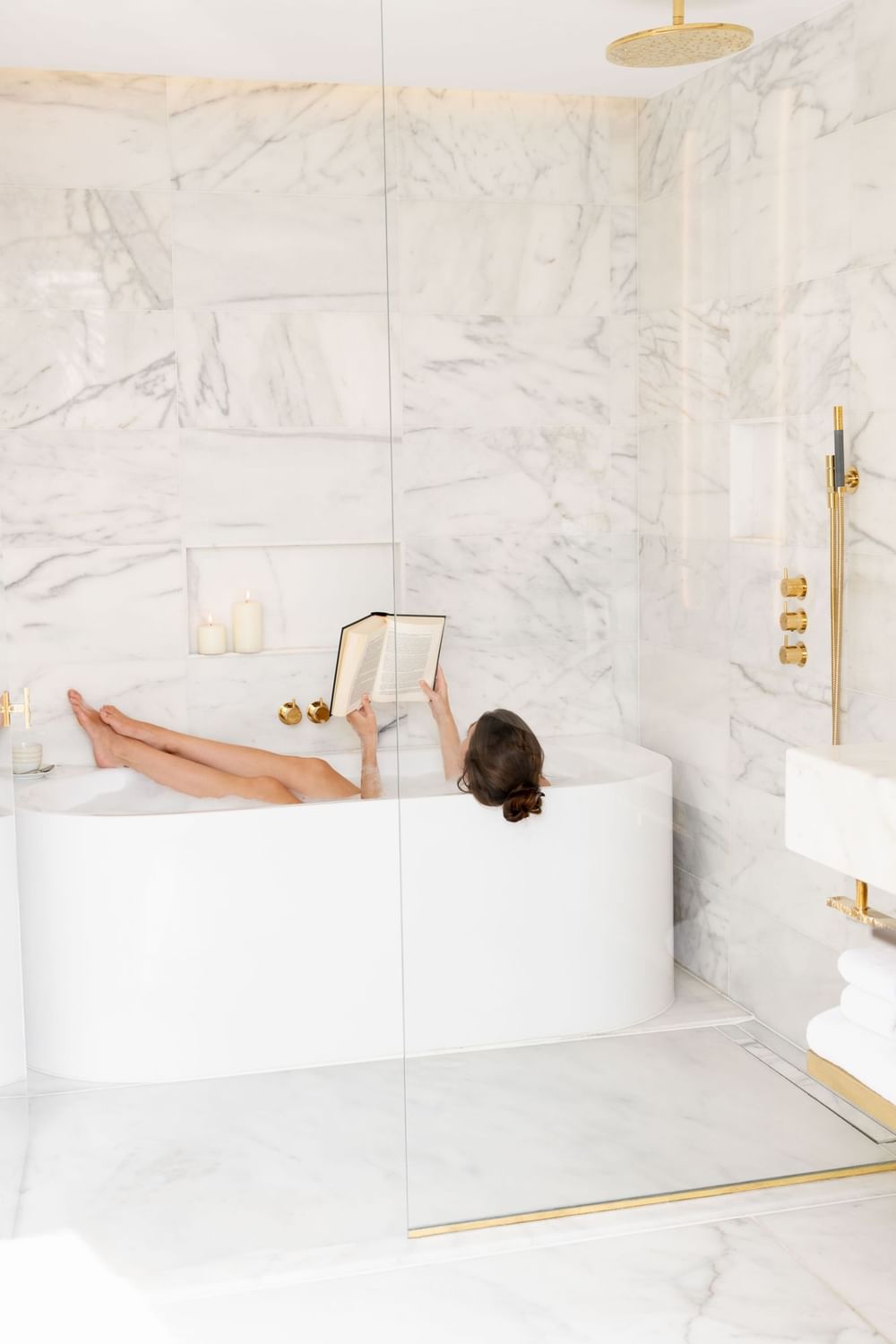 Woman relaxing in a luxurious marble bathroom tub while reading a book with candles at Almanac Barcelona