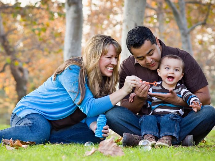 A family of three with a toddler blowing bubbles in a park.