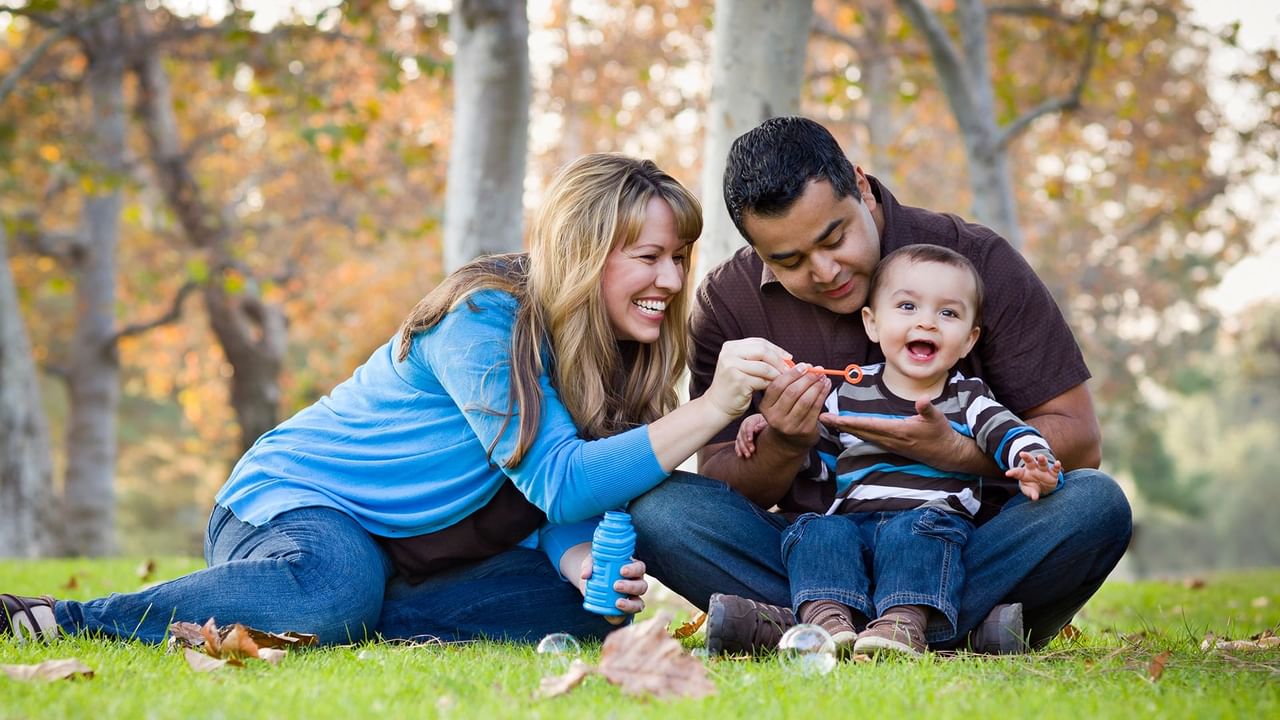 A family of three with a toddler blowing bubbles in a park.