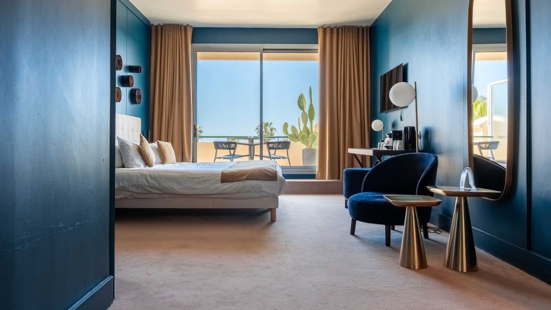 Chambre moderne aux murs bleus avec lit double, fauteuil en velours et terrasse ensoleillée avec vue sur la mer