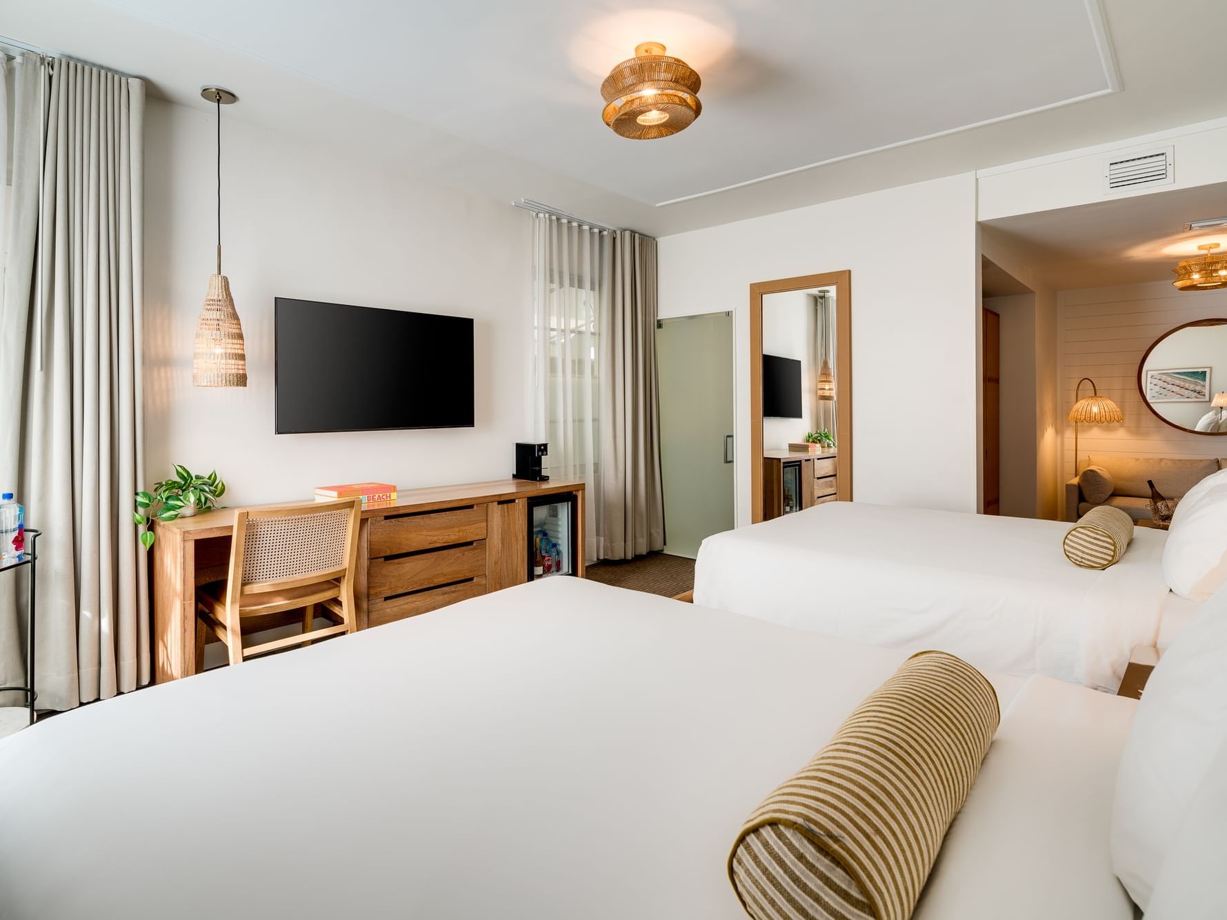 Bright and beach-chic hotel room featuring two Queen beds with crisp white linens and striped bolster pillows. The space includes a flat-screen TV mounted above a wooden console, a desk with a woven chair, pendant lighting, and a full-length mirror. Natur