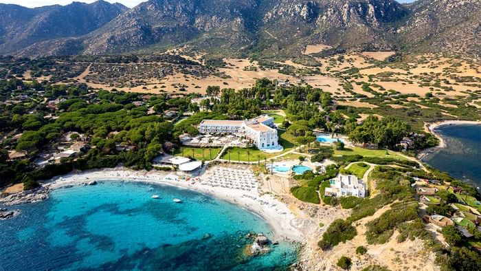 Aerial view of Falkensteiner Resort Capo Boi with sea, beach umbrellas, and mountains in the background