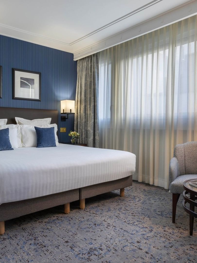 Large bed by a blue pinstripe wall under framed art with a gray armchair in a suite at Warwick Hotels & Resorts