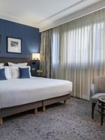 Large bed by a blue pinstripe wall under framed art with a gray armchair in a suite at Warwick Hotels & Resorts