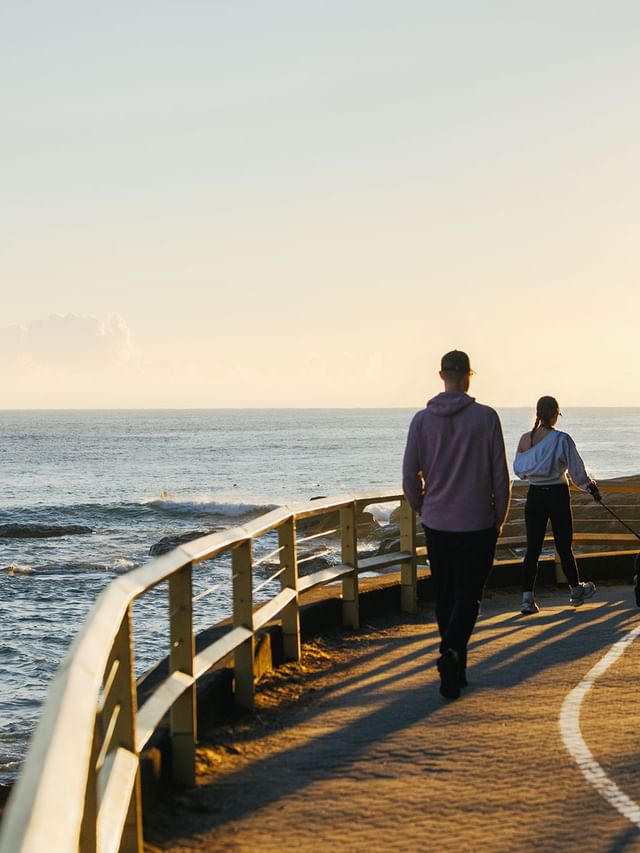 People walking a dog on a paved path by the ocean under a clear sky near a wooden fence at the scenic Elysium Manly