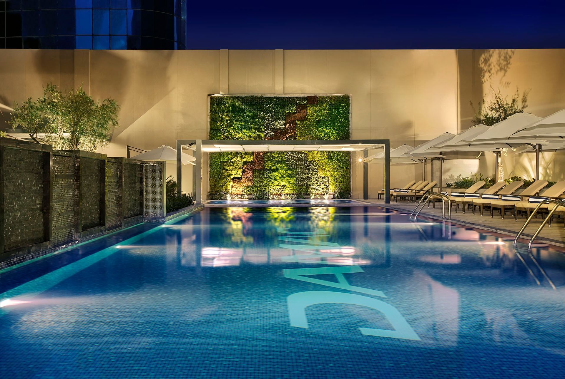 Luxurious poolside with loungers, ambient lighting, and a green wall at twilight at DAMAC Maison Cour Jardin
