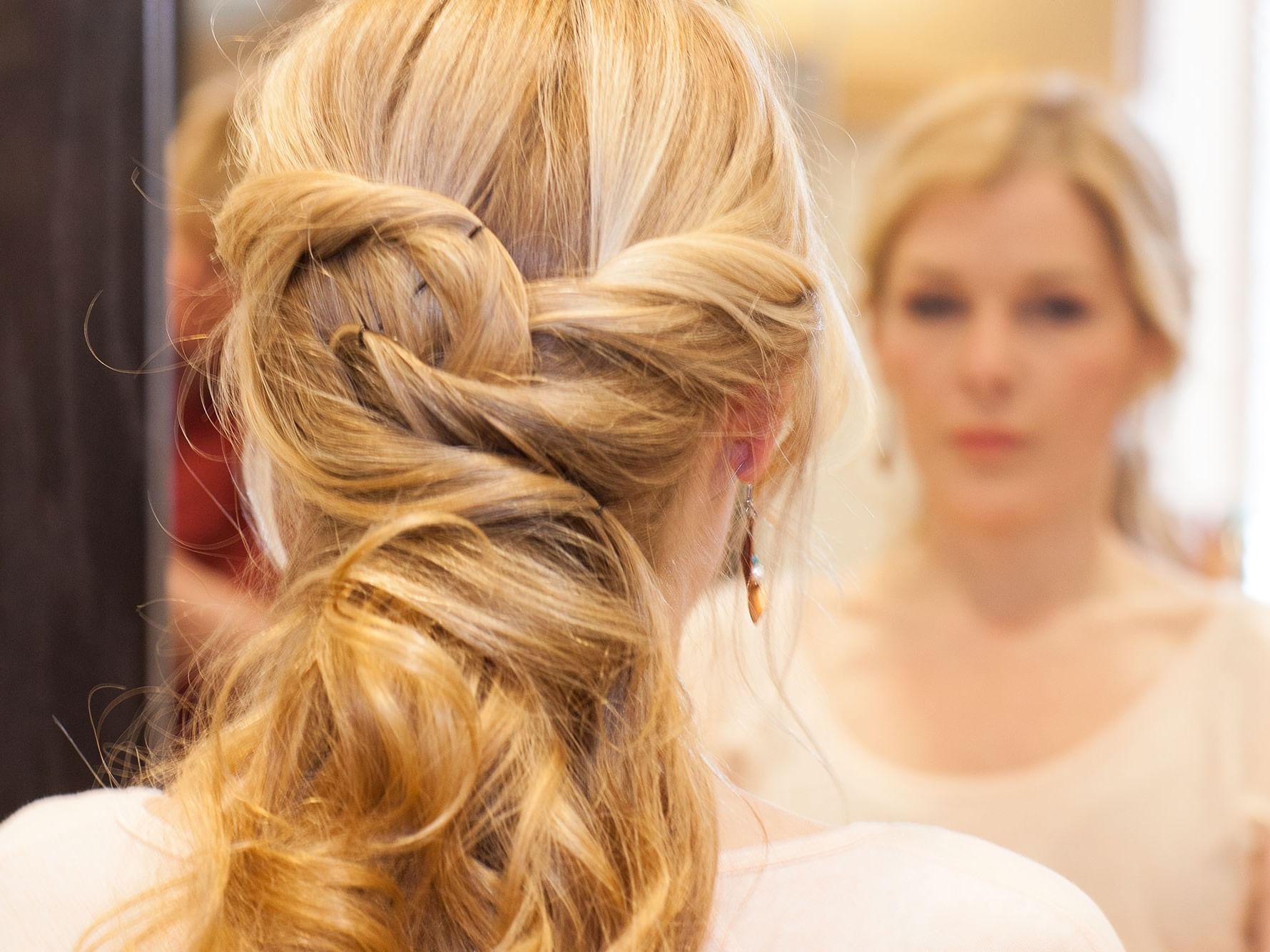 A woman with blonde hair styled in a braid looks at herself in a mirror.