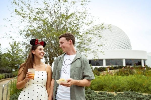 A young man and woman in Disney attire smile at one another with Spaceship Earth behind them at EPCOT.