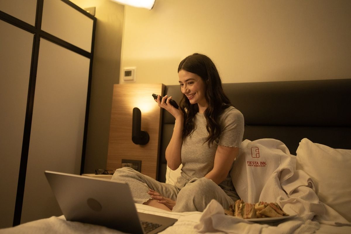 Una mujer sonríe hablando por teléfono mientras descansa en la cama con laptop y servicio a la habitación en Fiesta Inn