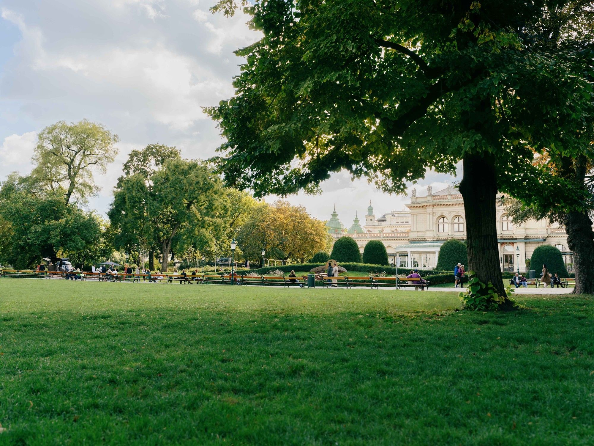 View across the wide lawns of Vienna’s Stadtpark with large shady trees, walking paths, and the Kursalon in the background