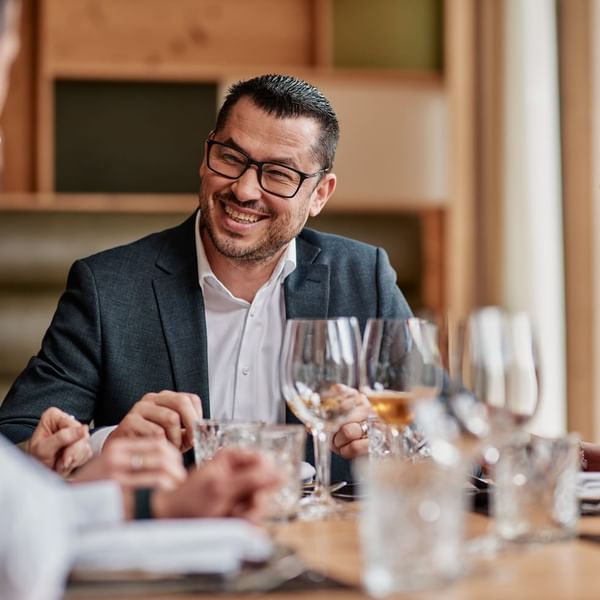 Mann mit Brille, Anzug und Krawatte sitzt am Tisch mit Wein und Wassergläsern und lächelt.