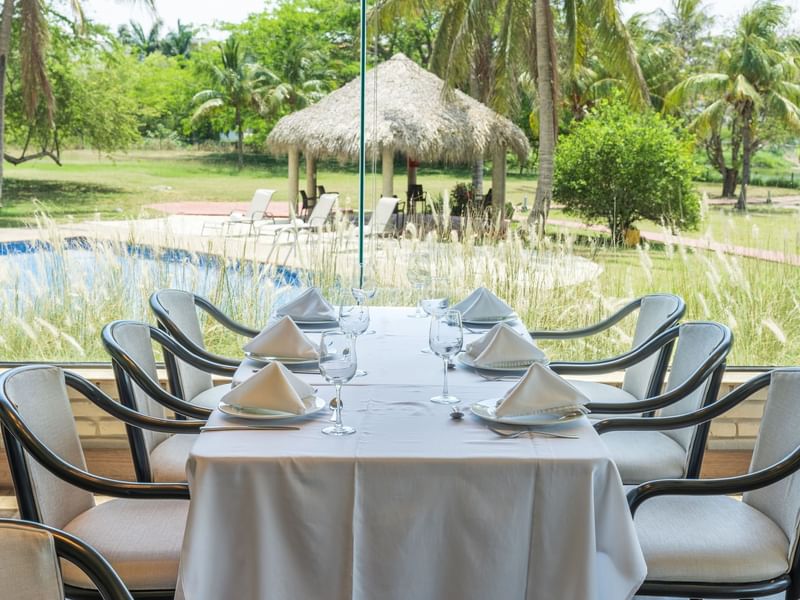 Dining table set with glassware, overlooking tropical garden with pool and thatched-roof gazebo at Fiesta Inn