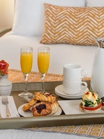 Breakfast tray with pastries and orange juice by tan pillows on a white bed in In-Room Dining at Warwick Melrose Dallas