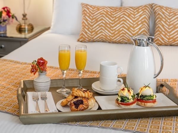 Breakfast tray with pastries and orange juice by tan pillows on a white bed in In-Room Dining at Warwick Melrose Dallas