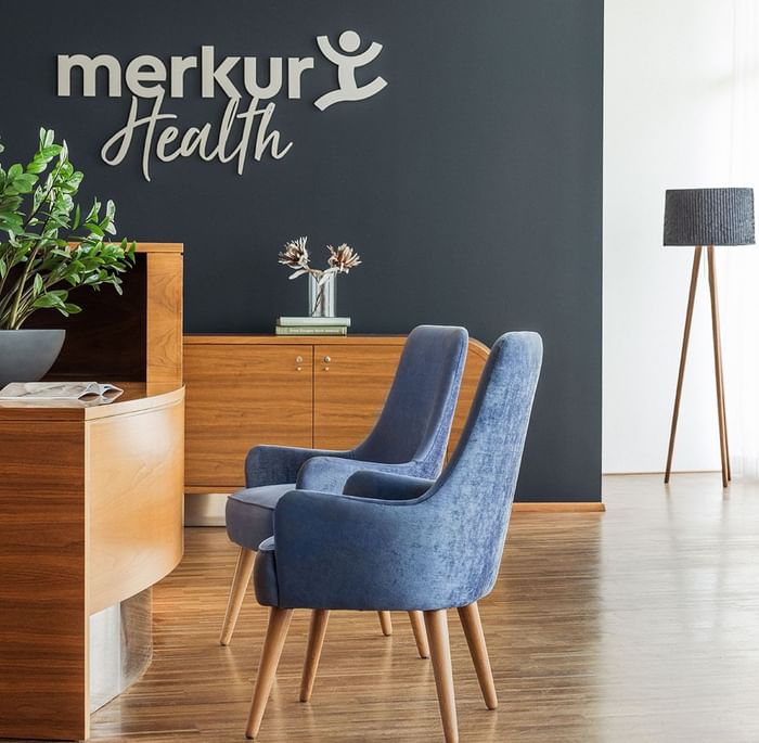 Sala d'attesa moderna con sedie blu e grigia, scrivania in legno, parete firmata Merkur Health e lampada.