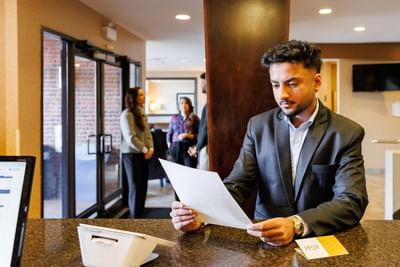 Businessman reading a document at the front counter check-in desk with hotel guests behind at Rodd Royalty
