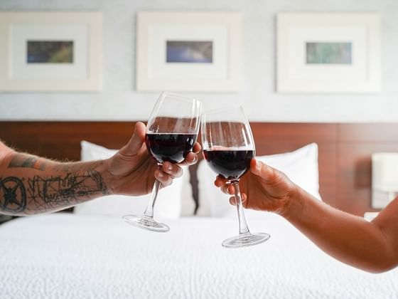Two hands holding glasses of red wine in a romantic toast above a plush white bed with pillows at Rodd Moncton