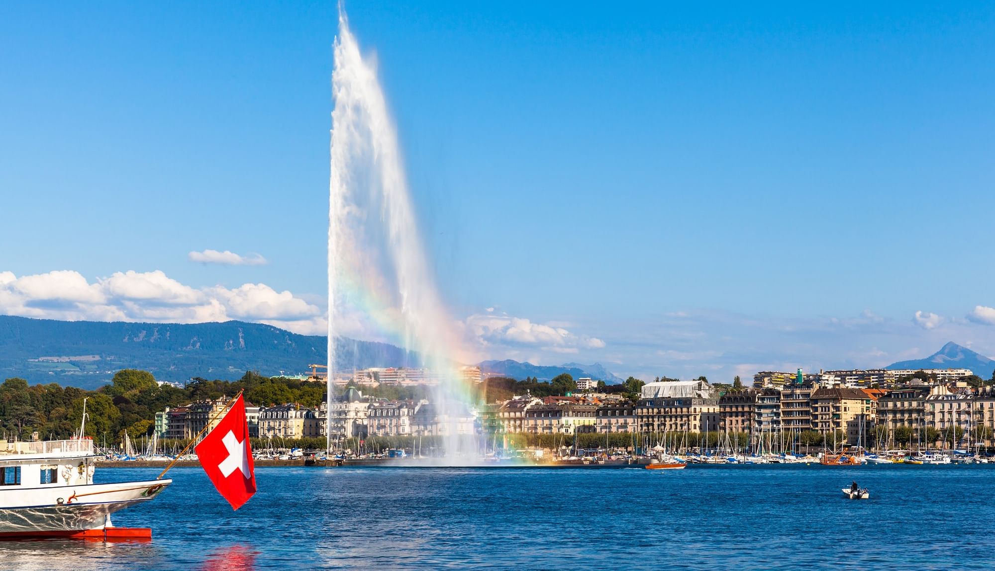 Water fountain Jet d'Eau with a rainbow by a boat and city buildings under a blue sky near Warwick Geneva