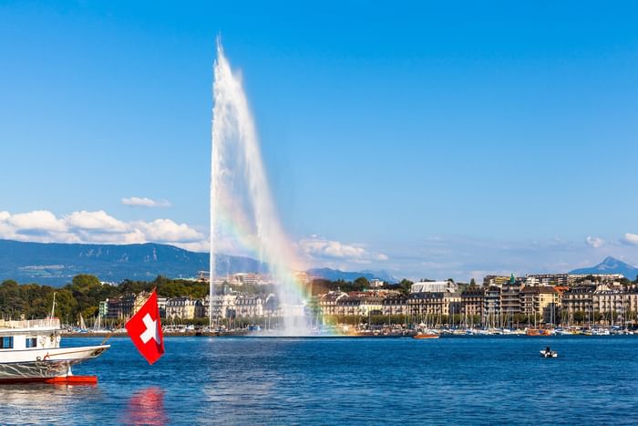 Water fountain Jet d'Eau with a rainbow by a boat and city buildings under a blue sky near Warwick Geneva