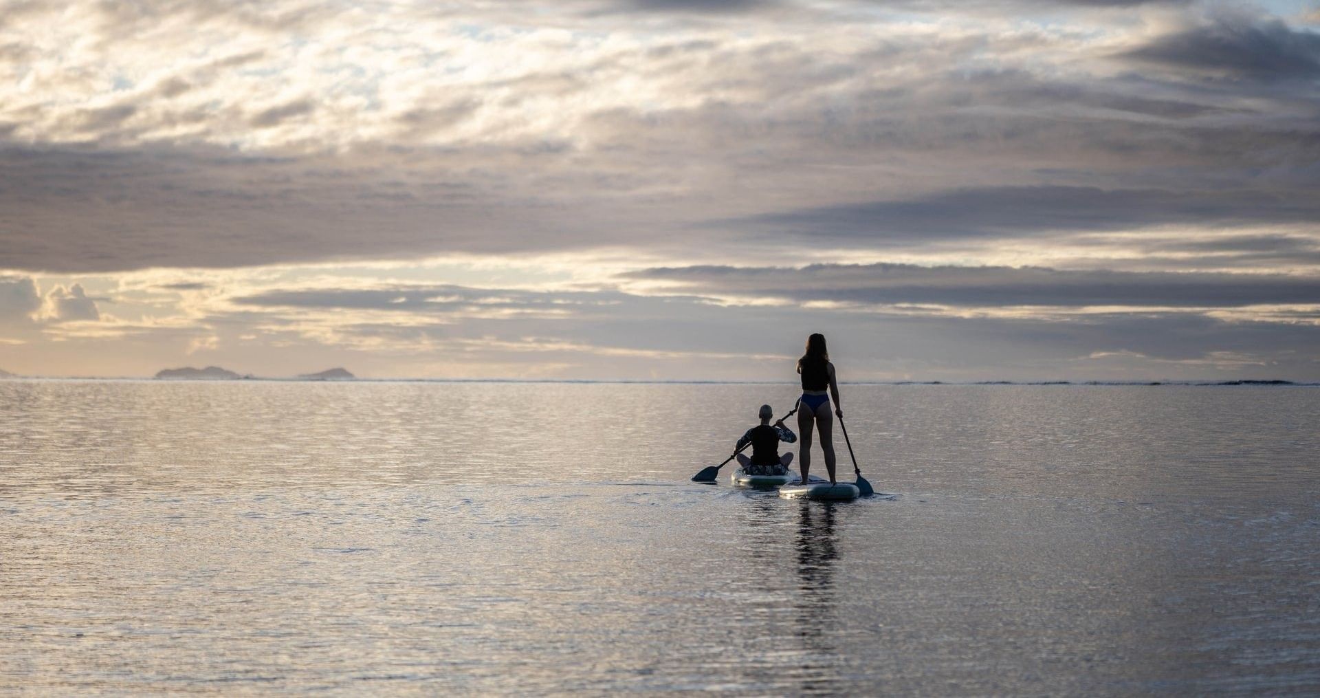 Two guests on paddleboards by a calm sea under a soft cloudy sunset near Warwick Fiji Resort and Spa