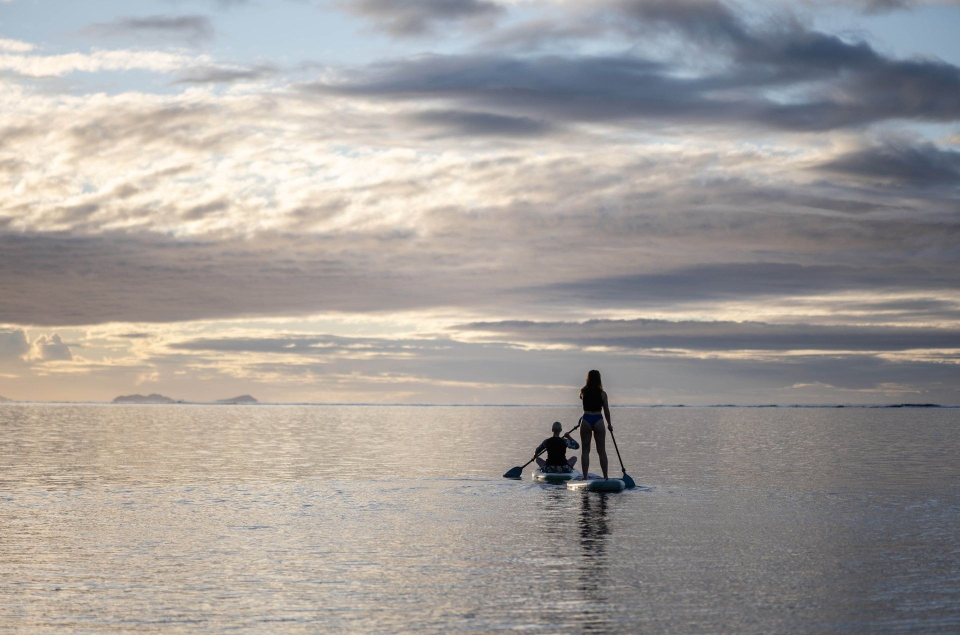 Two guests on paddleboards by a calm sea under a soft cloudy sunset near Warwick Fiji Resort and Spa