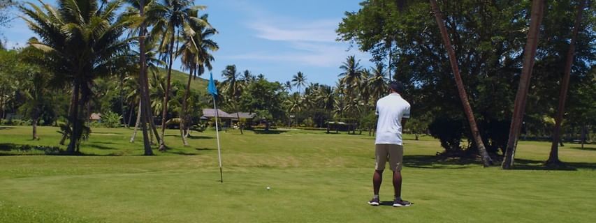 Man golfing on a green by a blue flag under the shade of leafy tropical trees at The Naviti Resort - Fiji