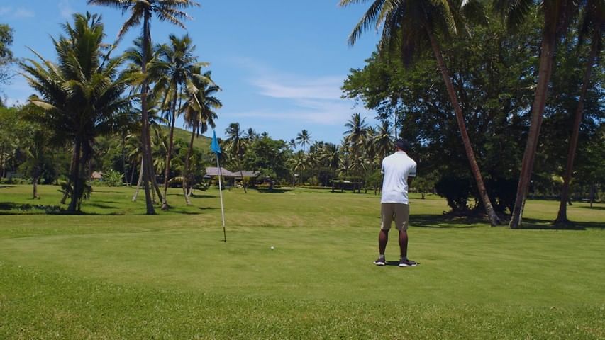 Man golfing on a green by a blue flag under the shade of leafy tropical trees at The Naviti Resort - Fiji