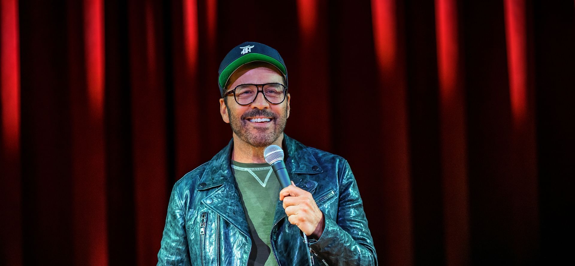 Actor and comedian Jeremy Piven wearing leather jacket and cap holding microphone on stage backdrop at The Brook