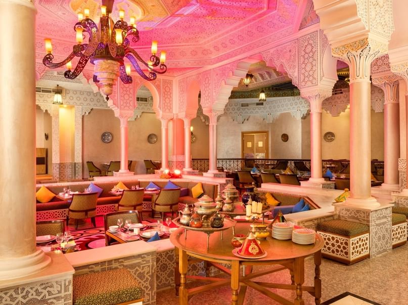 Elegant restaurant with ornate decor, cushioned seating, and a large chandelier, illuminated with pink lights.