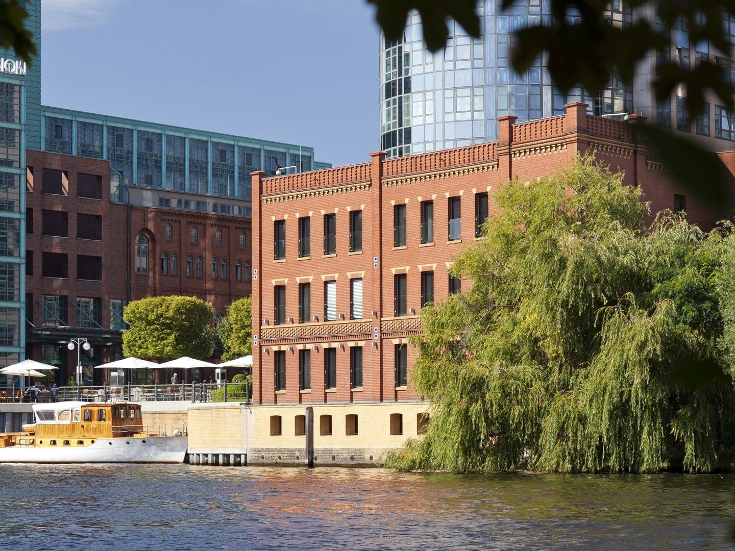 Ein Boot, das an einem roten Backsteingebäude angedockt ist, mit Bäumen und einer Außenterrasse mit Sonnenschirmen.